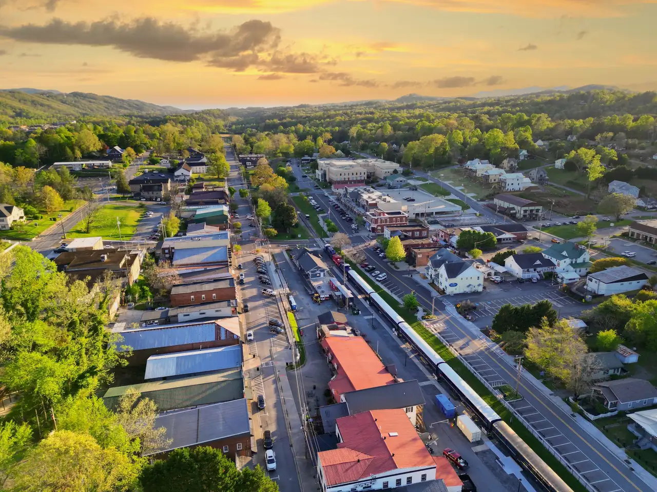 Aerial view at sunset of a small town grid with a long passenger train parked beside a depot, forested hills under an orange sky