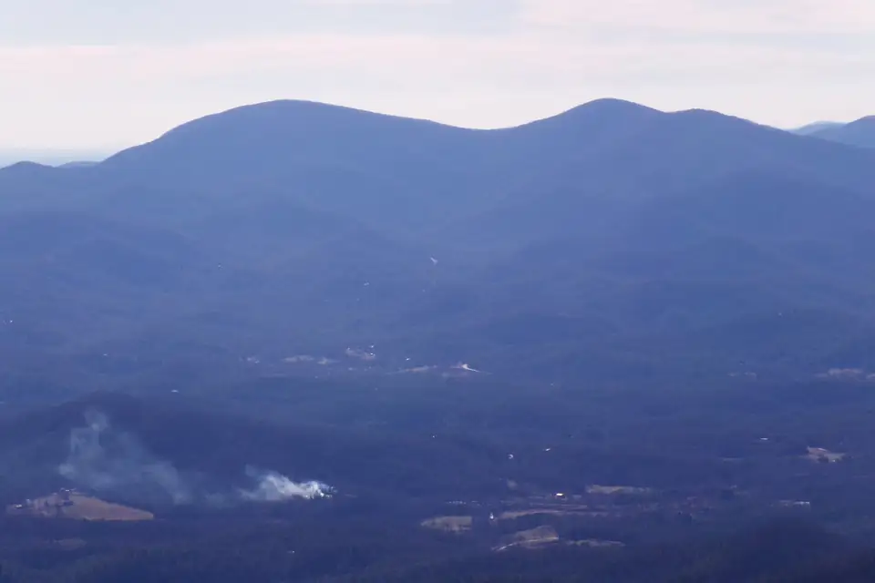 Hazy blue layered mountain ridges with a double-humped peak, farm clearings and drifting smoke in the valley below