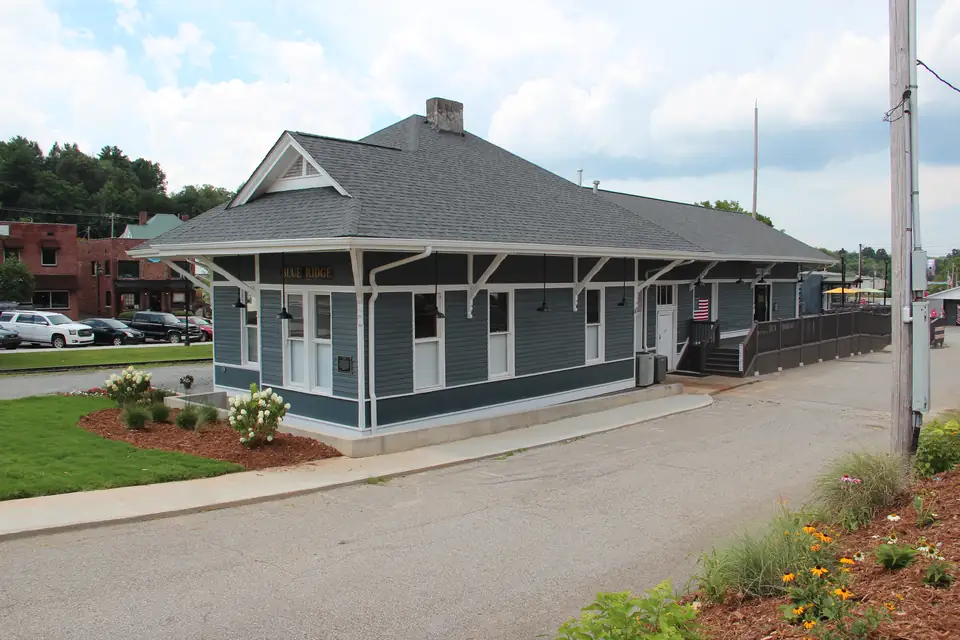 Gray-blue wooden railroad depot with white trim, gabled roof and a 'BLUE RIDGE' sign, flower beds and paved lot in front