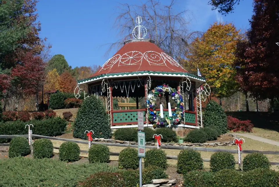 Red-roofed gazebo outlined with white light strands and a large wreath, candy-cane stakes with red bows along a rail fence