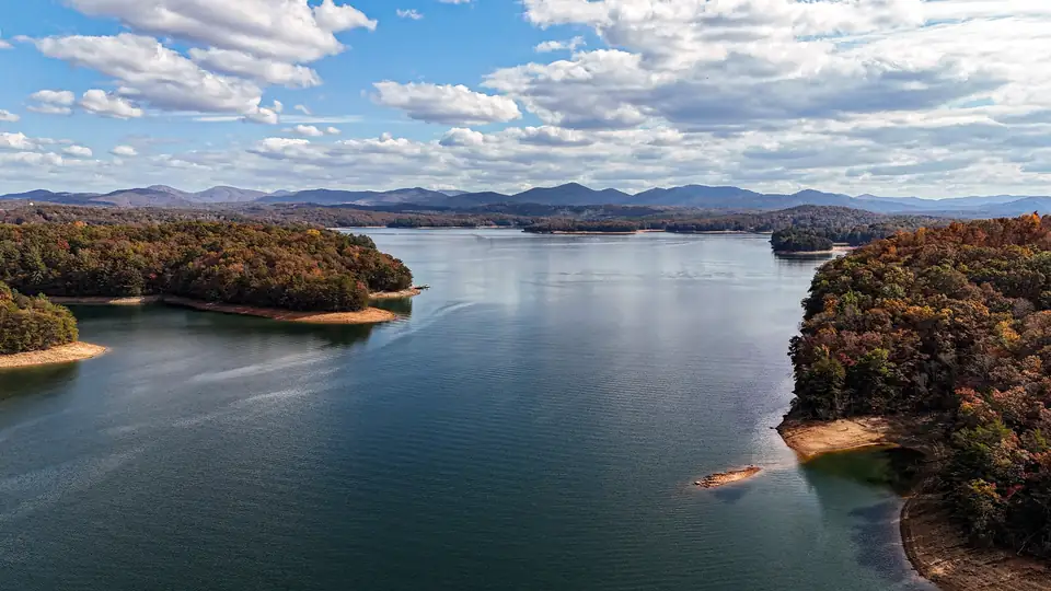 Aerial view of a wide blue lake between wooded points in orange and brown fall color, layered mountains and white clouds beyond