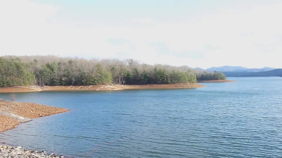 Rippled blue lake with a reddish exposed shoreline, a pine-covered point and distant blue mountains under a pale sky