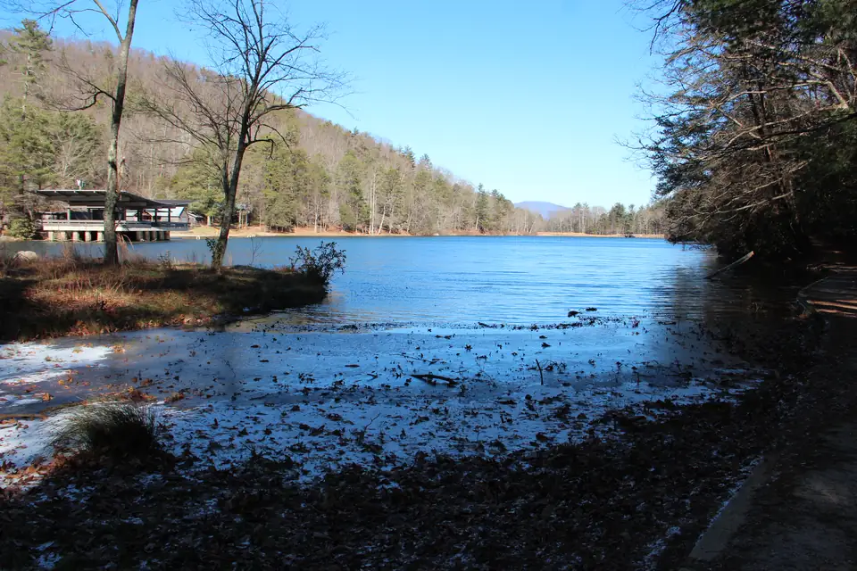 Ice-edged shoreline with fallen leaves in the foreground, a blue lake, a lakeside pavilion and bare and evergreen trees on hills