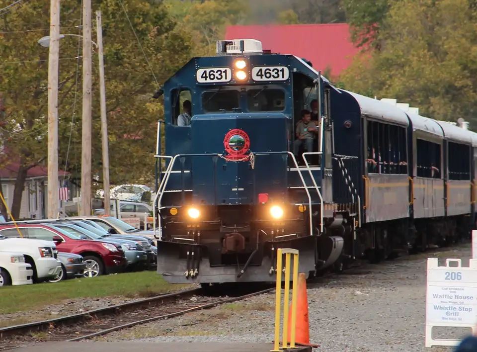 Blue diesel locomotive numbered 4631 with headlights on, pulling open-window passenger coaches past parked cars and an orange cone