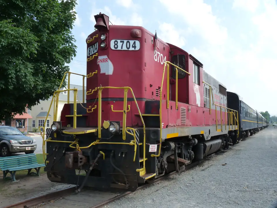 Red diesel locomotive numbered 8704 with yellow handrails coupled to blue passenger coaches, a green bench and tree at left