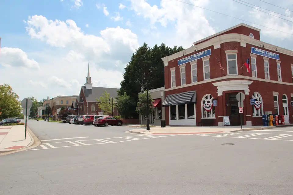 Red-brick corner building with striped awning and patriotic bunting, a crosswalk, parked cars and a white church steeple down the street
