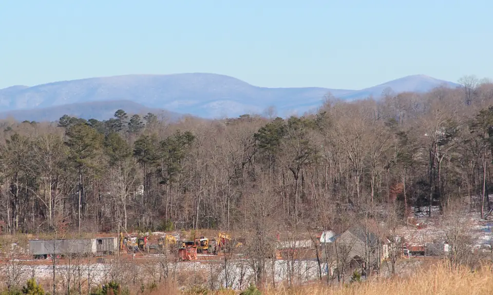Snow-dusted blue mountain ridges above a bare winter forest, with trucks and equipment in a lot and snow patches below