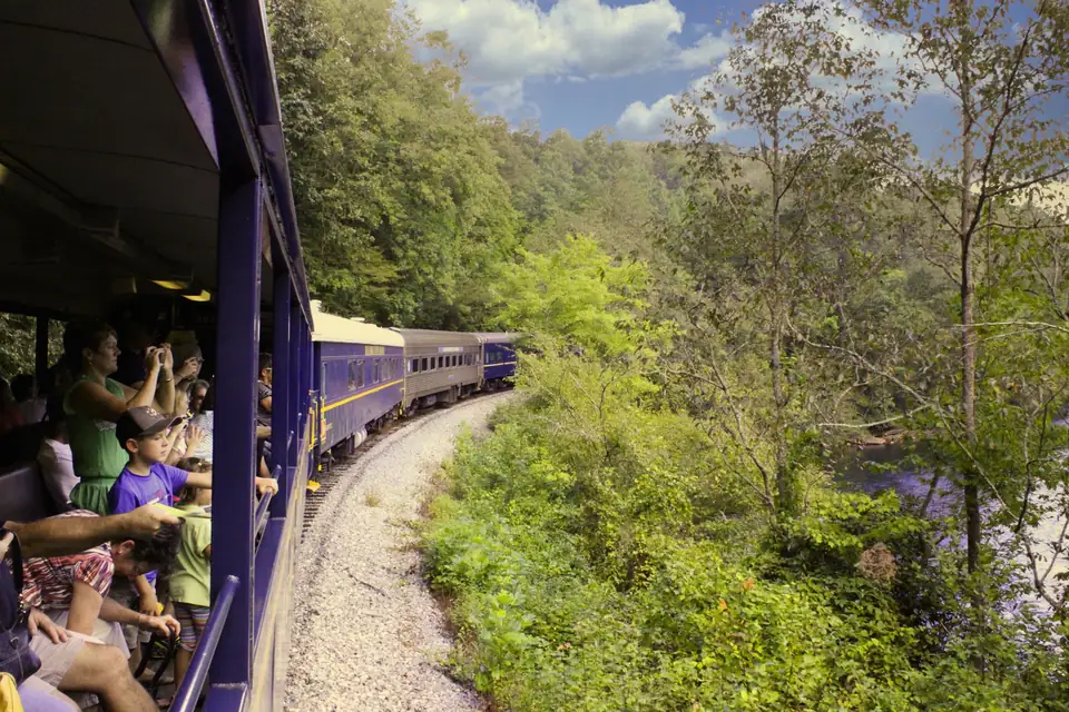 Passengers in an open-air rail car photograph the view as blue and silver coaches curve ahead through green woods beside a river