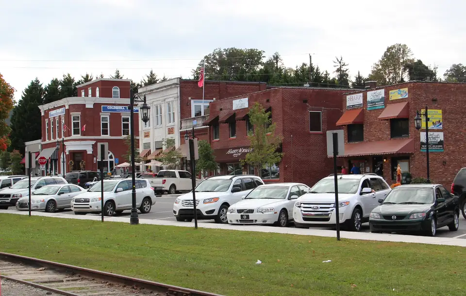Row of two-story brick storefronts with awnings and lanterns, white cars parked in front, a rail track and lawn in the foreground