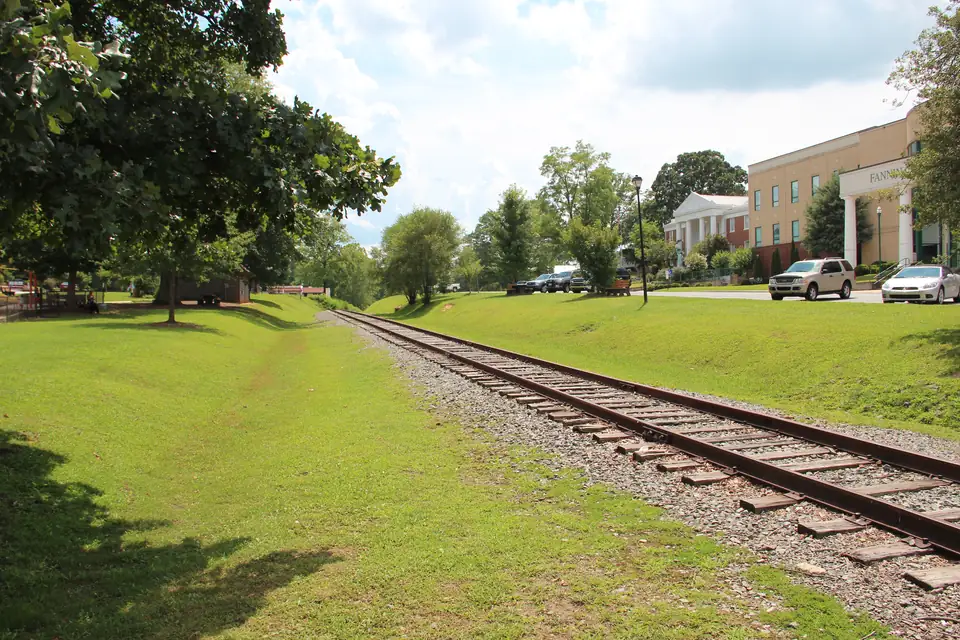 Single railroad track running through a grassy park strip beside a cream government building with columns and parked cars